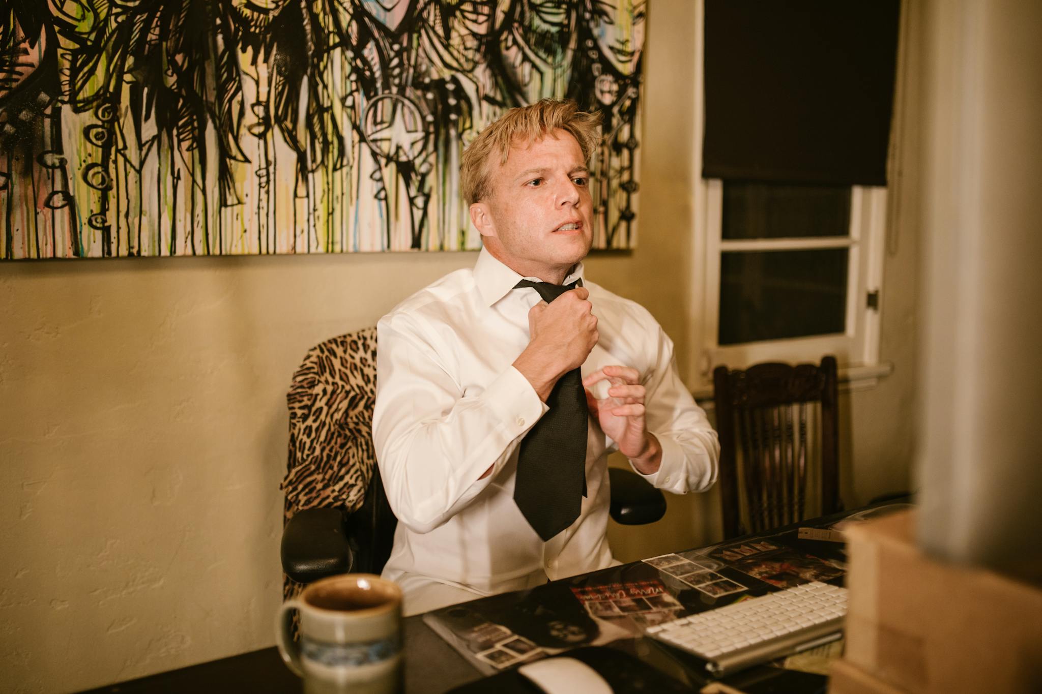 An adult man adjusts his tie in a home office, ready for a virtual meeting, with a cup of coffee on the desk.
